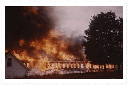 Building on fire with smoke and flames billowing out, surrounded by trees and a house.